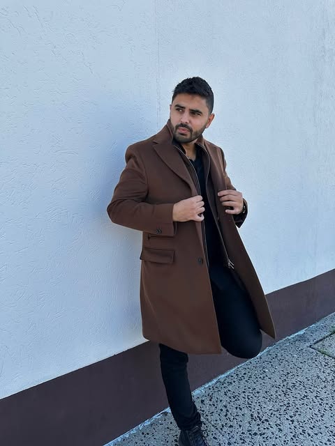 Alex Katz in a brown overcoat, leaning on a white wall.