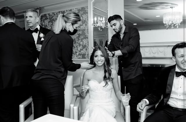 Black-and-white wedding-day photo: a smiling bride in her gown, with long loose waves, while two stylists finish her hair.